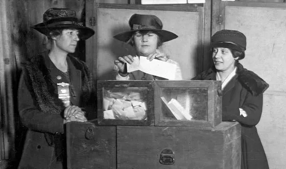 Description: black-and-white photo of three women in early 20th Century clothes, at a wooden ballot box. The woman in the center folds her ballot to place in the box. 